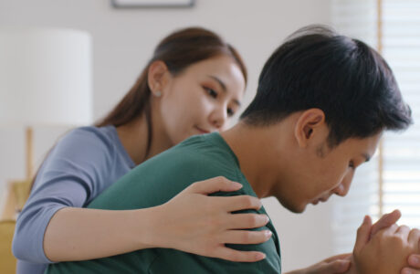 A young Asian female puts her arm around a young Asian man, who has his hands clasped and appears to be in grief